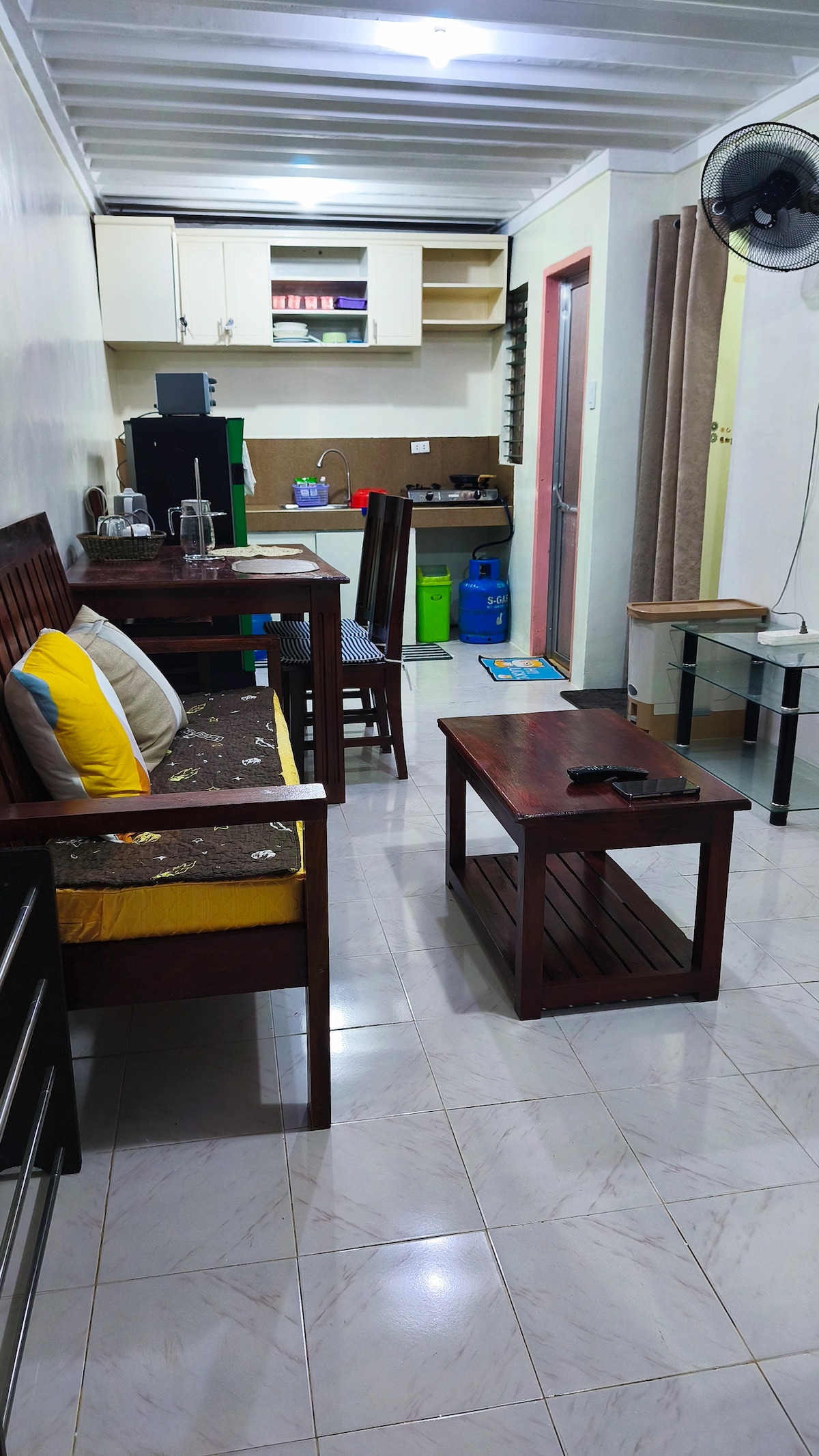 A living area features a small wooden coffee table and two wooden chairs, complemented by a sofa with yellow cushions. A compact kitchen is visible in the background, equipped with basic amenities. A floor fan and glass side table are included, enhancing the functional layout.
