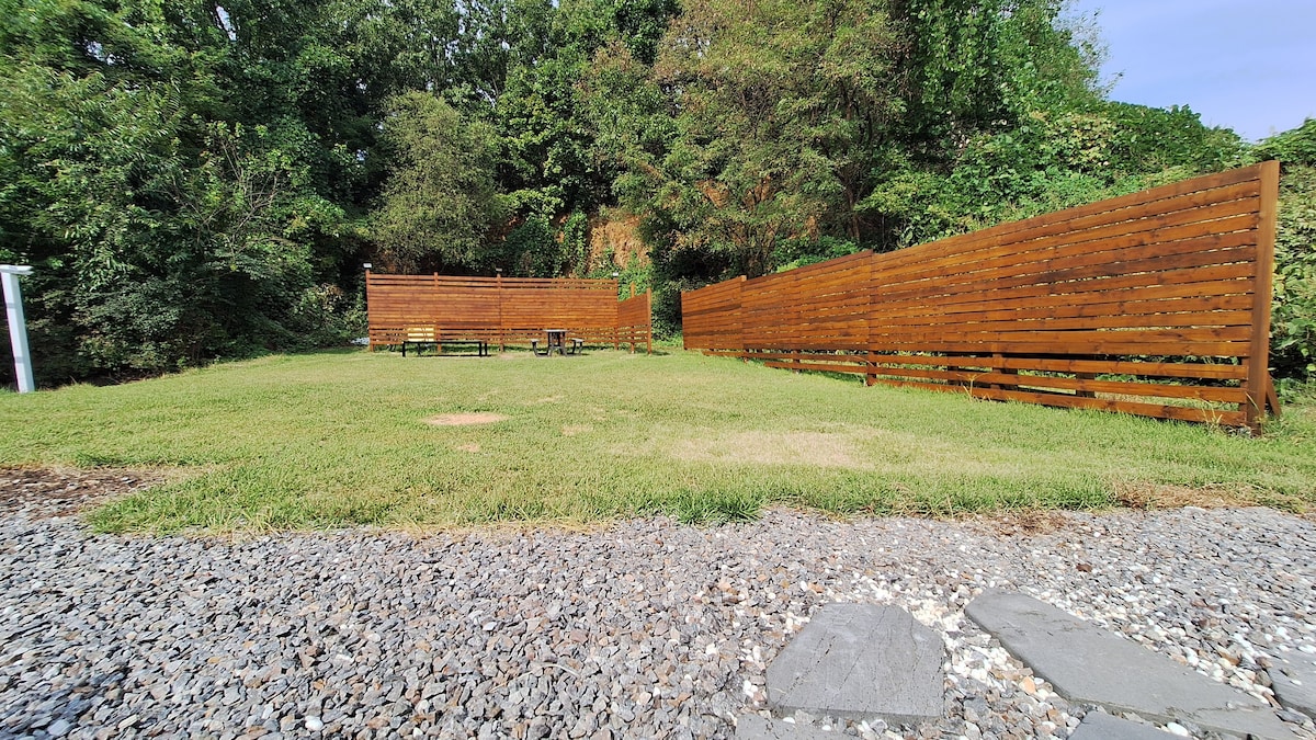A spacious outdoor area features a well-maintained lawn surrounded by wooden walls, offering privacy. Paved stones and gravel comprise the ground, providing access to the grassy space, which is dotted with a few seating options, suitable for relaxation or gathering.