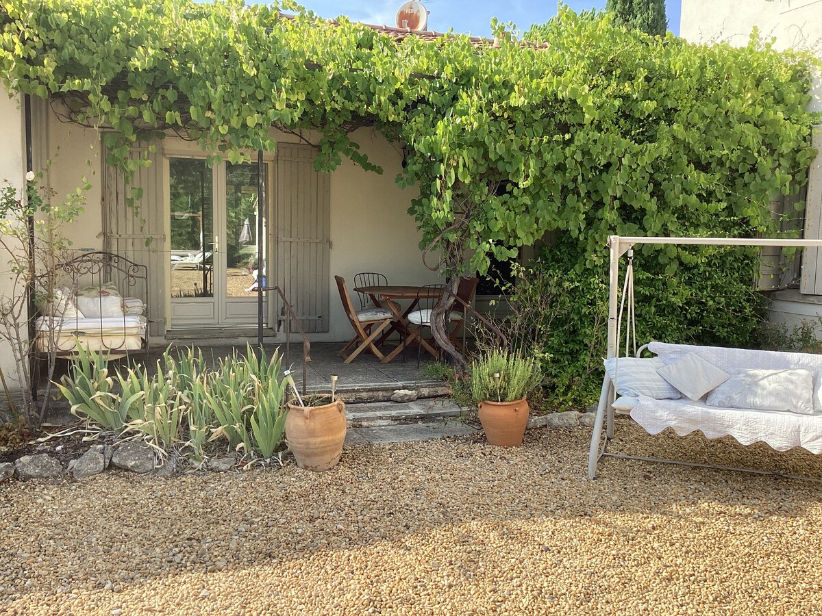 A charming outdoor space features a gravel area with potted plants and a swing chair. A small dining table is set for two under a leafy canopy, while light streams in from the open French doors leading inside the house.