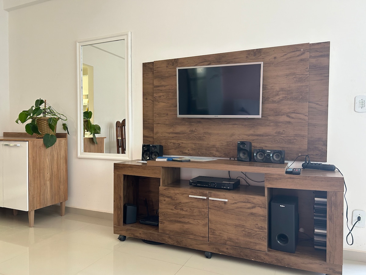 A living area features a wooden entertainment unit with a flat-screen TV, speakers, and a media player. A mirror is mounted on the wall, and a green potted plant adds a touch of nature. Light-colored flooring complements the neutral tones of the furniture.