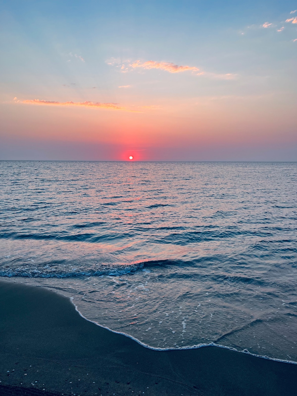 A serene view of the horizon at sunset, where soft hues of pink and orange blend into the tranquil blue of the sea. Gentle waves can be seen lapping at the shore, creating a peaceful atmosphere.