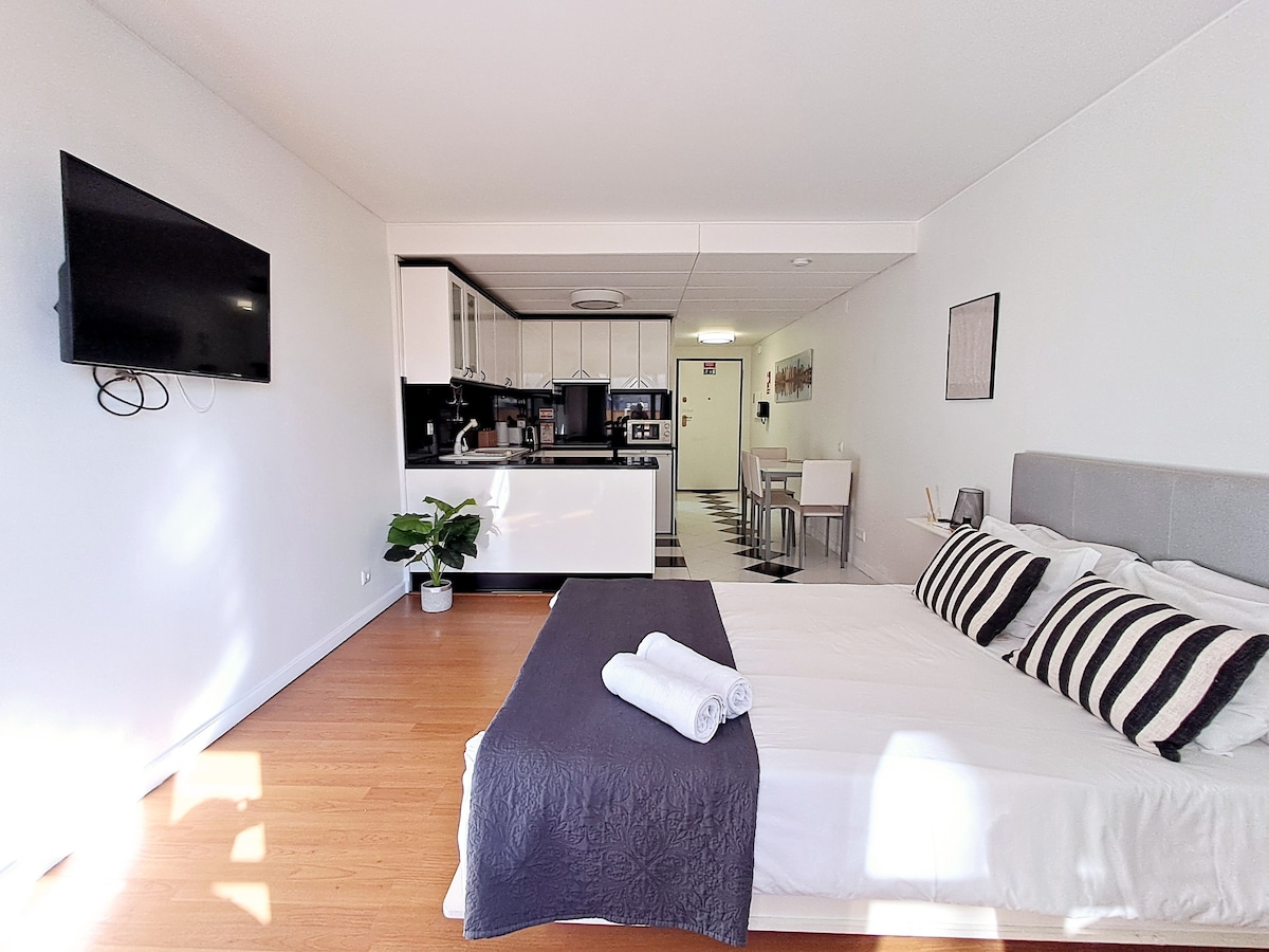 An open-plan studio is illuminated by natural light, featuring a cozy sleeping area with a spacious bed adorned with striped pillows. A dining table and kitchen are visible in the background, along with a television mounted on the wall. A small plant adds a touch of greenery.