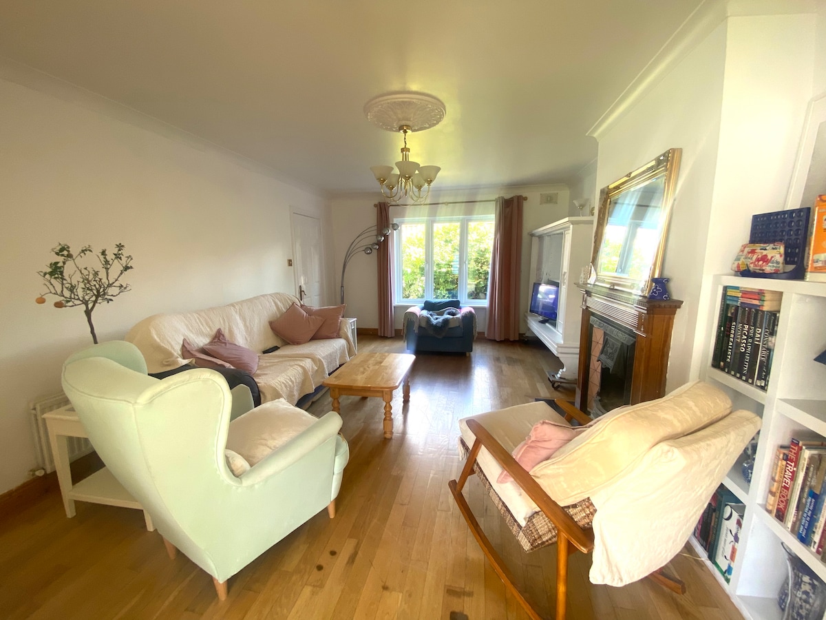 A bright living room features a mix of seating options, including a light green armchair and cream sofas. A wooden coffee table is positioned at the center, while natural light floods the space through large windows adorned with sheer curtains. Decorative items are seen on shelves.