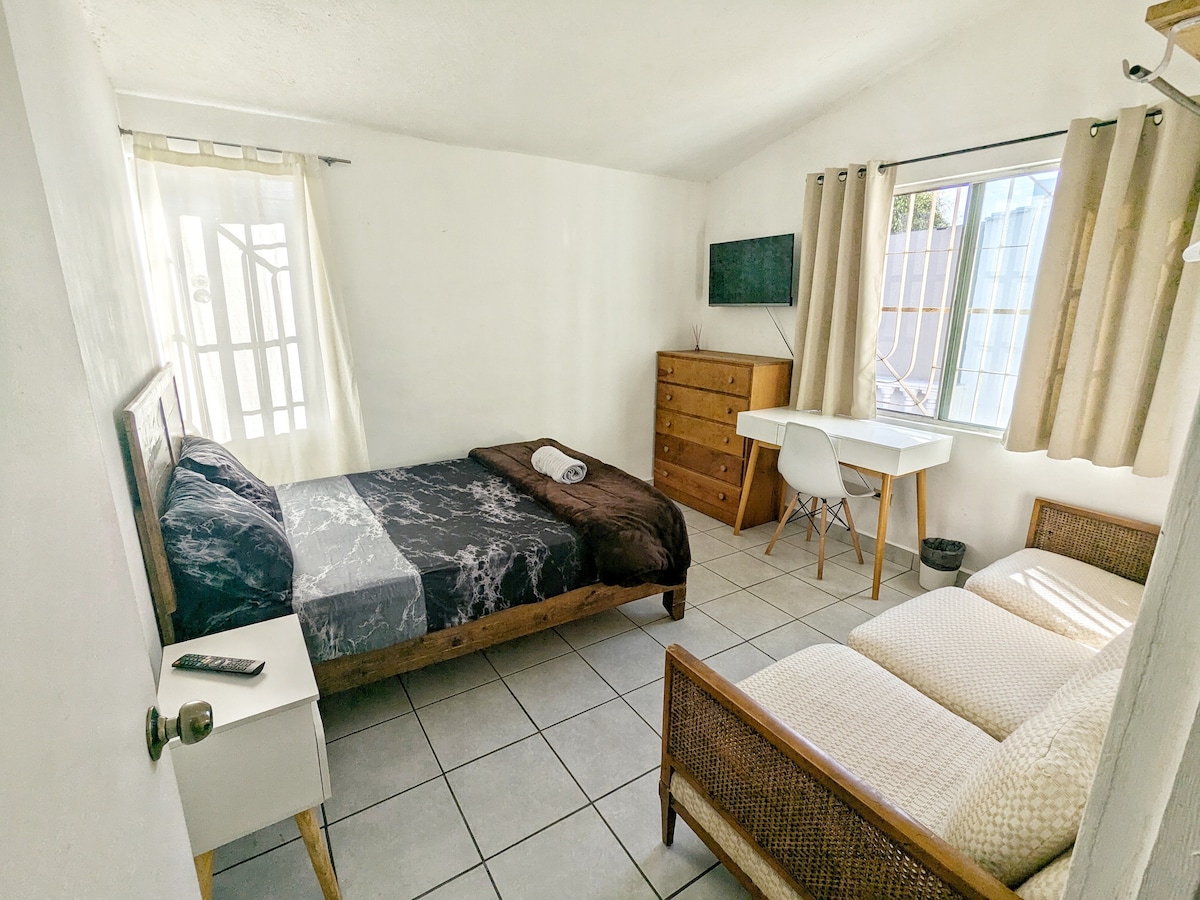 A bright room features a spacious bed with a dark blanket and a slim profile dresser. A cozy sofa provides extra seating, while a desk with a chair offers a workspace. Natural light enters through a window adorned with light curtains.