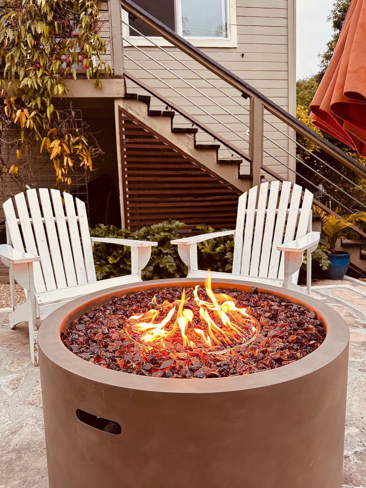 A circular fire pit is surrounded by two white Adirondack chairs, creating a cozy seating area. The flames flicker gently above the polished stones in the center. In the background, steps lead to the entrance of the house, framed by greenery.