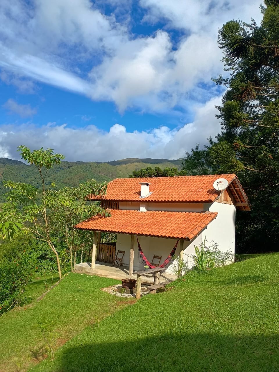 A charming chalet with a red-tiled roof is set against a backdrop of green mountains and a blue sky. A porch extends from the house, featuring a hammock and a small table, creating a serene outdoor space surrounded by lush grass.