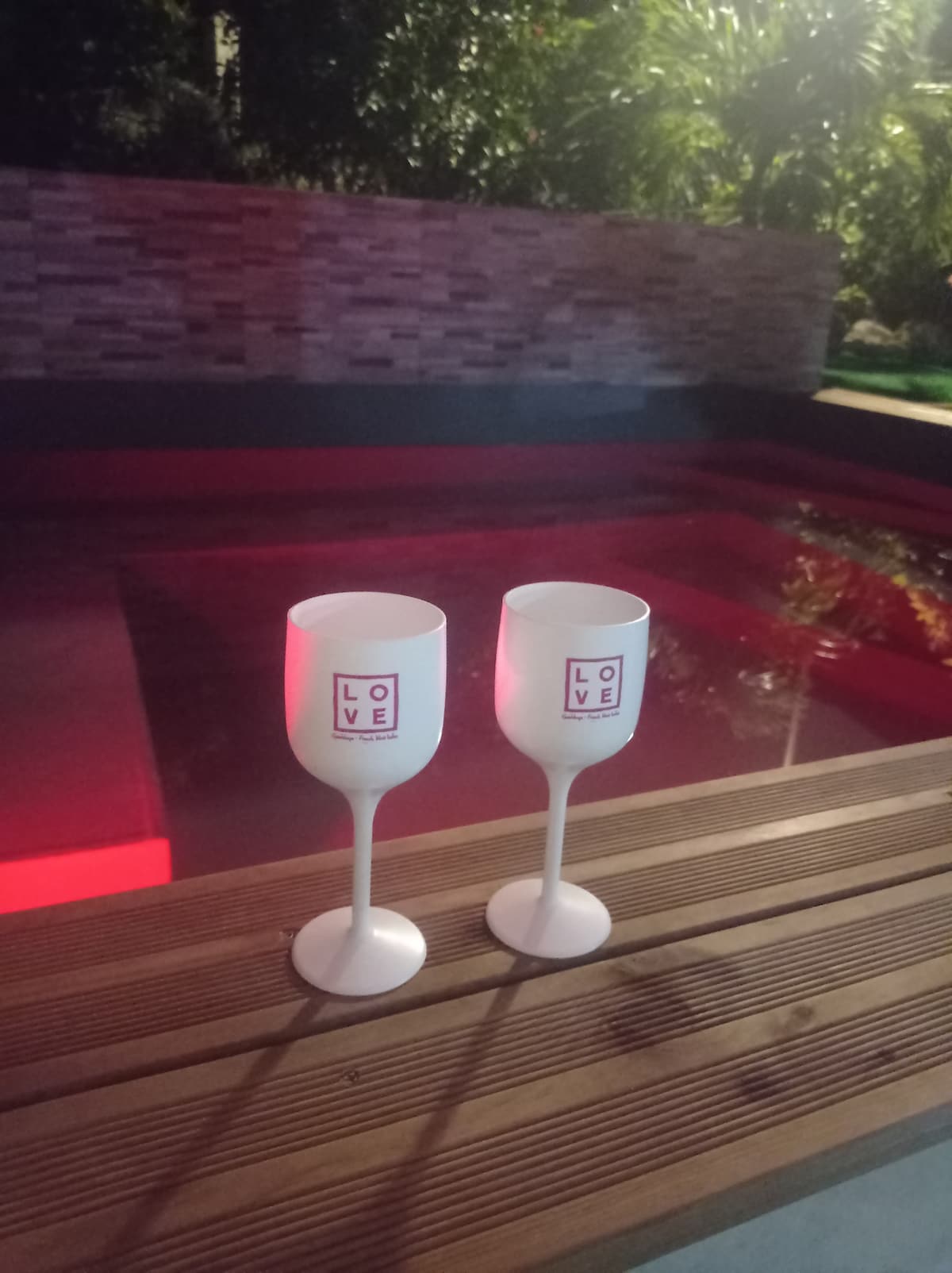 Two white glasses, labeled with the word 'LOVE,' stand on a wooden edge beside a pool. The pool reflects soft red lighting, creating a calm evening ambiance. Surroundings include lush greenery and stone wall features, enhancing a serene outdoor setting.
