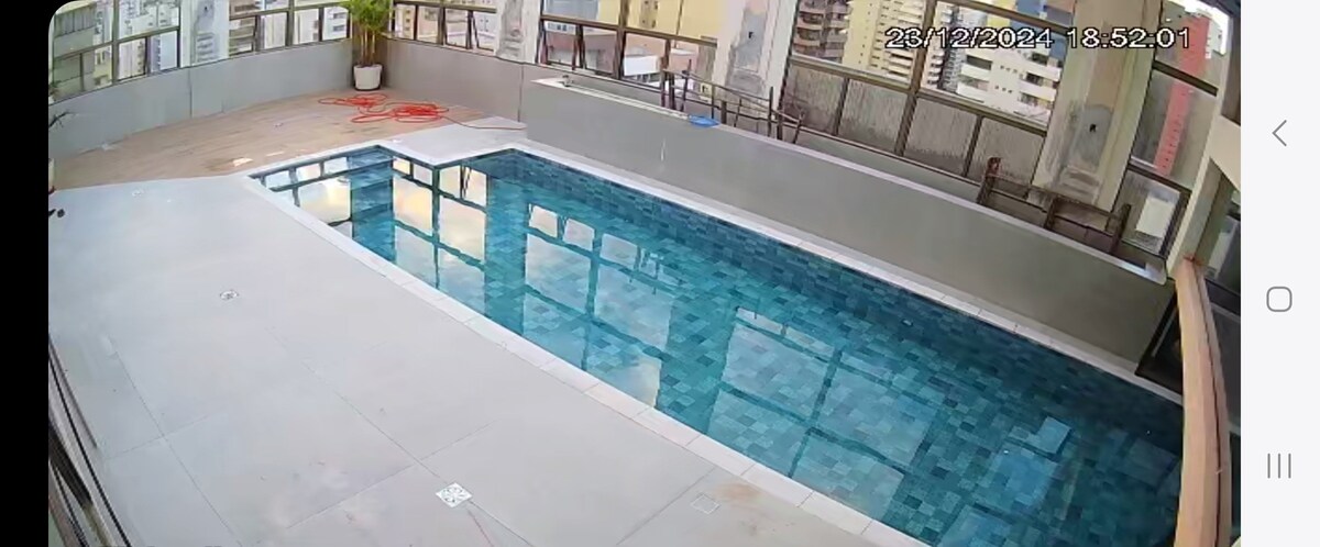 A rectangular swimming pool is visible, featuring a blue tiled bottom and clear water reflecting natural light. Surrounding the pool, a spacious deck area provides a place for relaxation, with glass railings offering views of the nearby cityscape. The setting appears inviting and open.