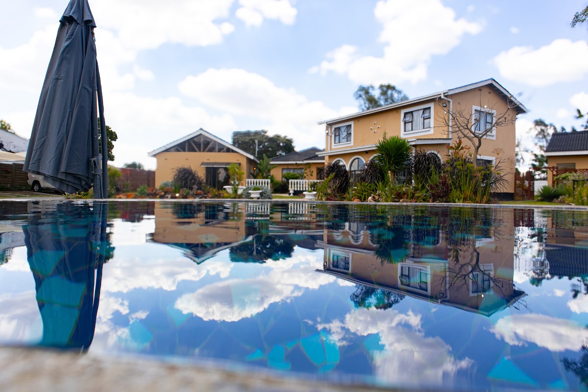 A tranquil swimming pool reflects the blue sky and fluffy clouds, framed by a well-manicured garden. The two-story house and adjacent structures are visible in the background, featuring a mix of light-colored walls and greenery.