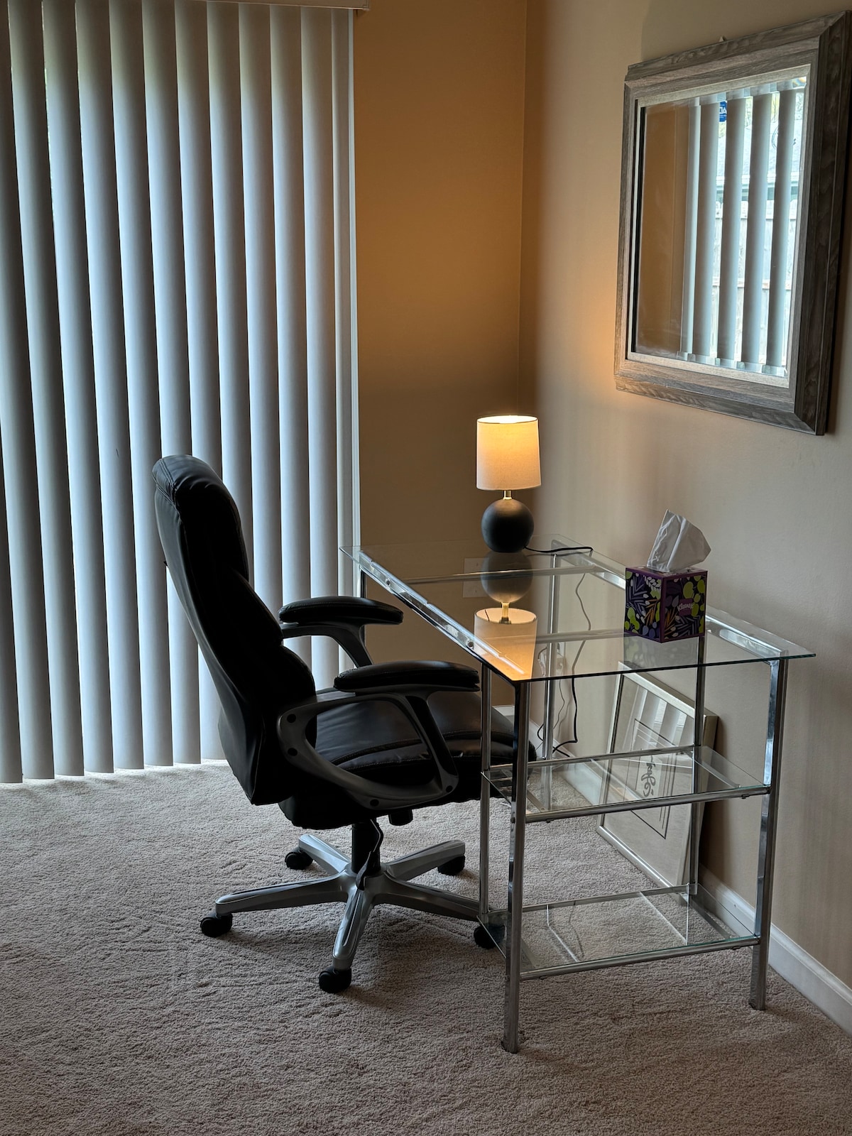 A workspace is provided featuring a clear glass desk paired with a comfortable black office chair. A lamp with a soft glow and a decorative tissue box are present on the desk. Vertical blinds allow natural light to filter into the room.