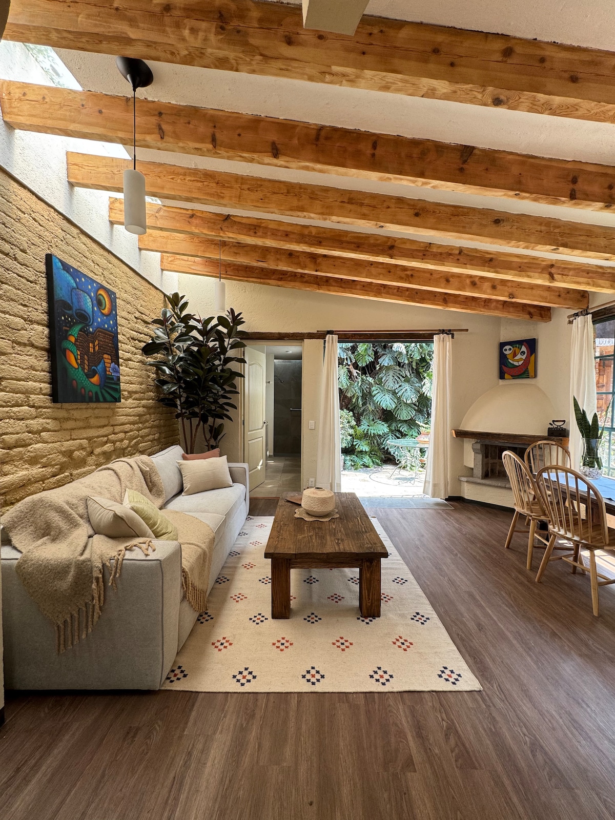 A spacious living area features a light-filled design with wooden beams overhead. A comfortable sofa is positioned beside a wooden coffee table, while two dining chairs are placed near a rustic dining table. Vibrant artwork adorns the walls, and greenery is visible through the windows.