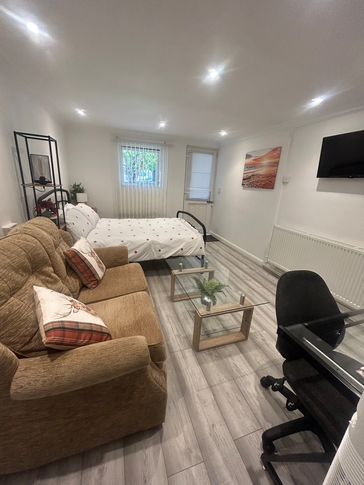 A comfortable living space features a spacious bed with white linens and decorative accents. A cozy couch is positioned nearby, accompanied by a glass coffee table. A desk with a chair is also present, while a window allows natural light to fill the room.