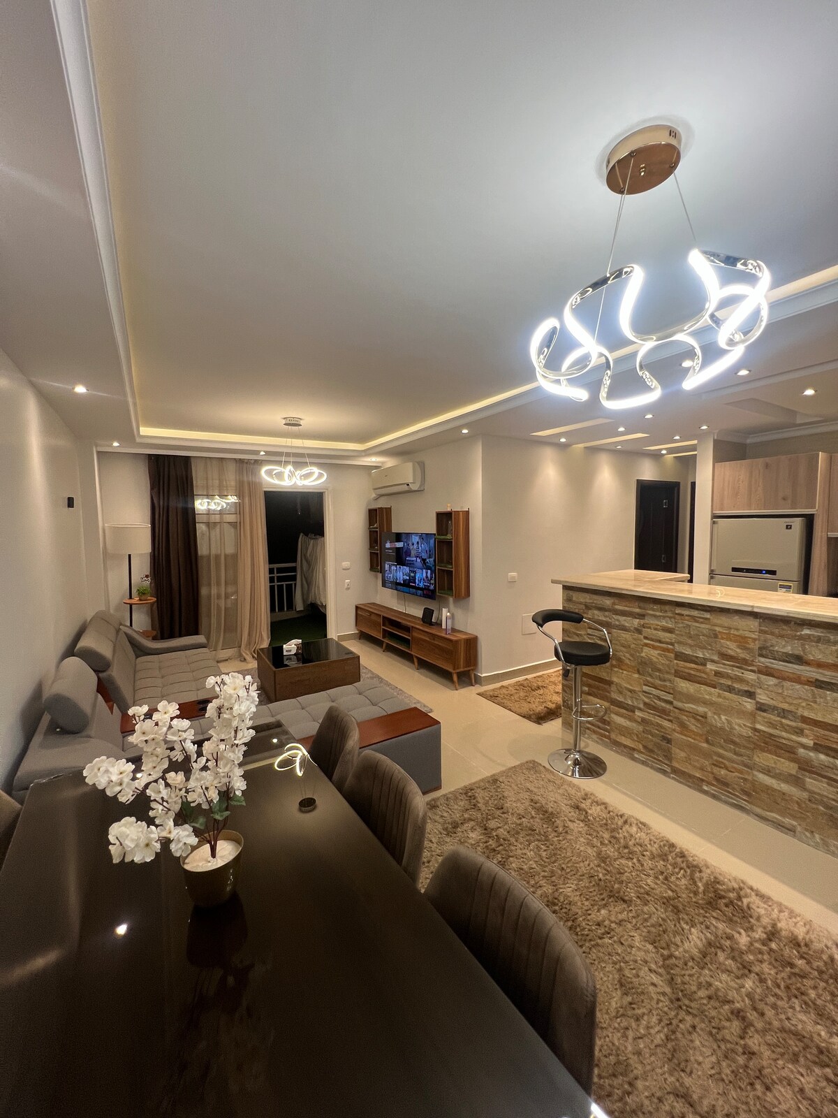 An inviting living area features a stylish dining table surrounded by upholstered chairs. A plush rug covers the floor, while soft lighting is provided by contemporary fixtures. The kitchen, visible in the background, includes a bar with high seating and modern appliances.