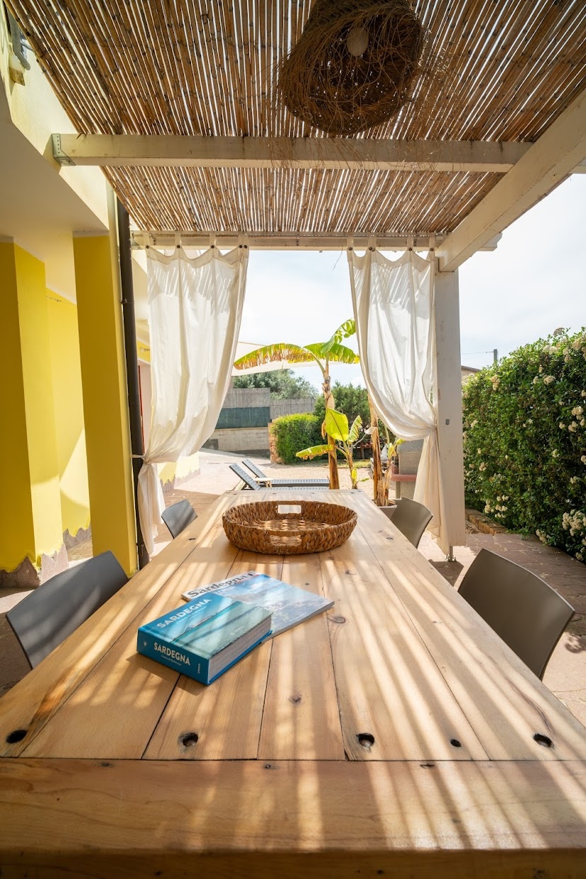 An outdoor dining area is highlighted by a large wooden table surrounded by chairs. A woven centerpiece sits atop the table, while sheer curtains provide soft light and shade. Potted plants and greenery are visible in the background, enhancing the outdoor setting.