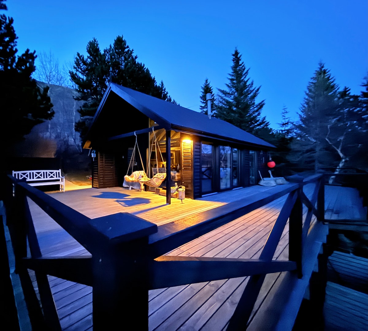 The exterior of the cottage is illuminated in the evening light, showcasing a spacious wooden deck with a swing chair. Tall trees surround the structure, and large glass doors provide a view into the cozy interior. A peaceful atmosphere is created by the natural setting.