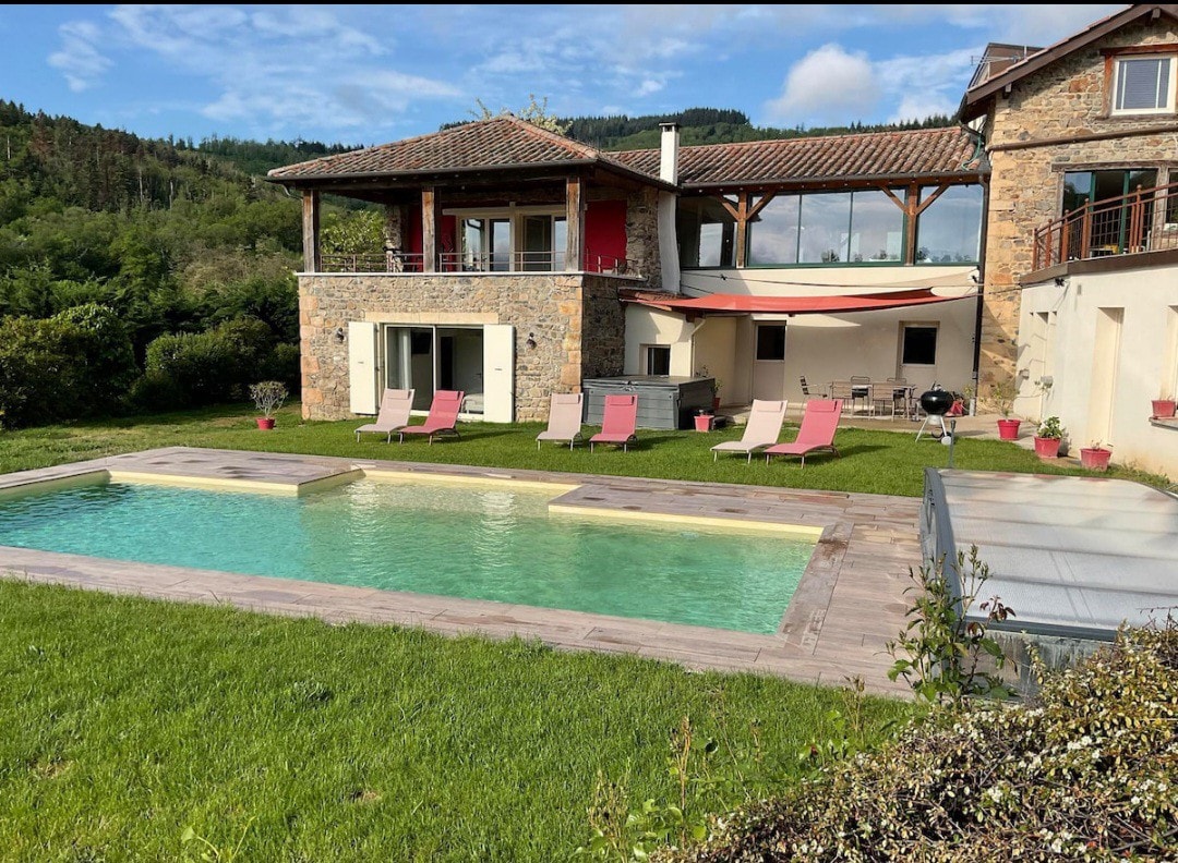 A serene outdoor space features a rectangular pool surrounded by lush green grass. Lounge chairs in various colors are positioned near the water. The two-story house, built with natural stone, displays an open design with large windows and a shaded terrace area, promoting relaxation.