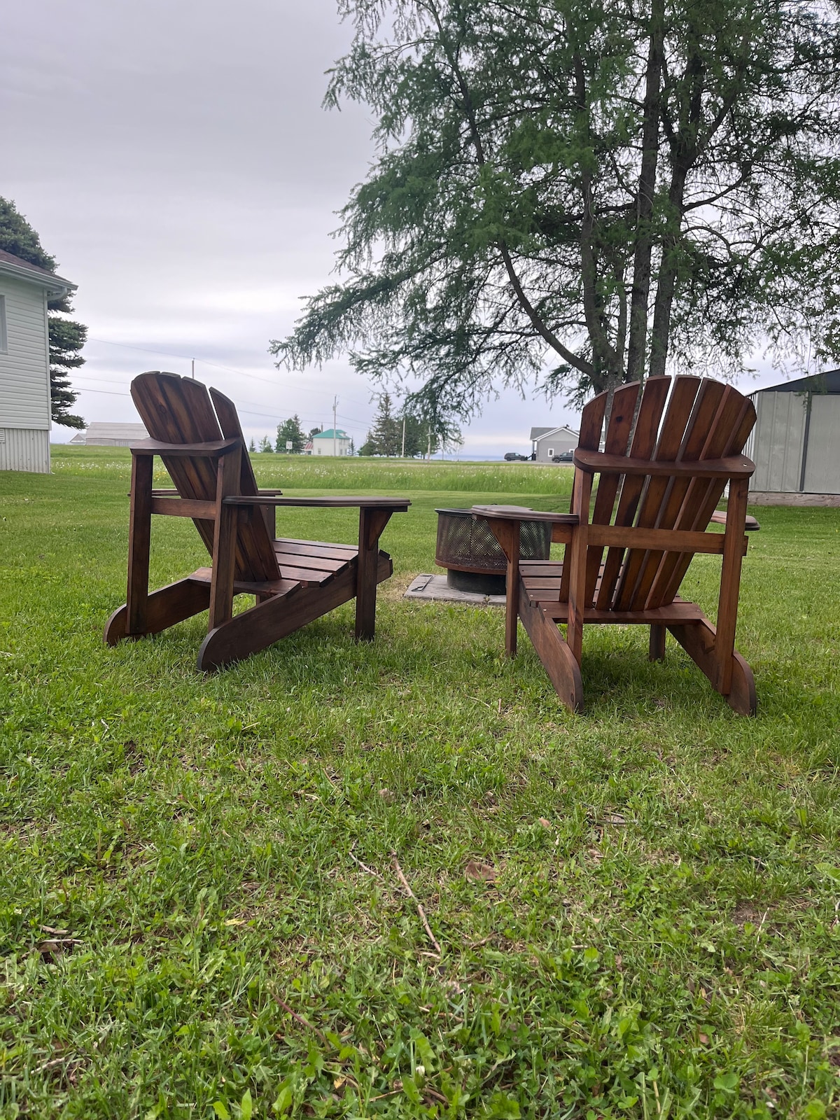 Two wooden Adirondack chairs are positioned in a grassy area, facing an outdoor fire pit. A large tree provides some shade nearby, and an open landscape extends in the background, offering a sense of tranquility.