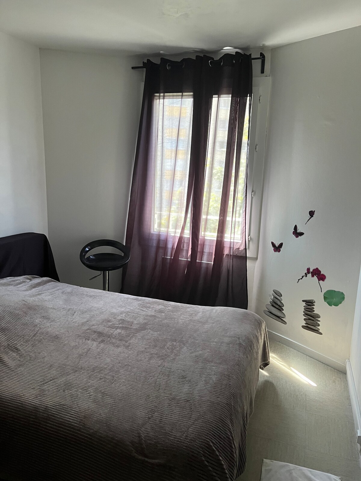 A cozy bedroom features a spacious bed covered with a soft, neutral-colored blanket. Natural light filters through a window dressed with sheer black curtains. A decorative wall showcases butterfly decals, adding a touch of charm, while a small, modern chair is positioned near the window.