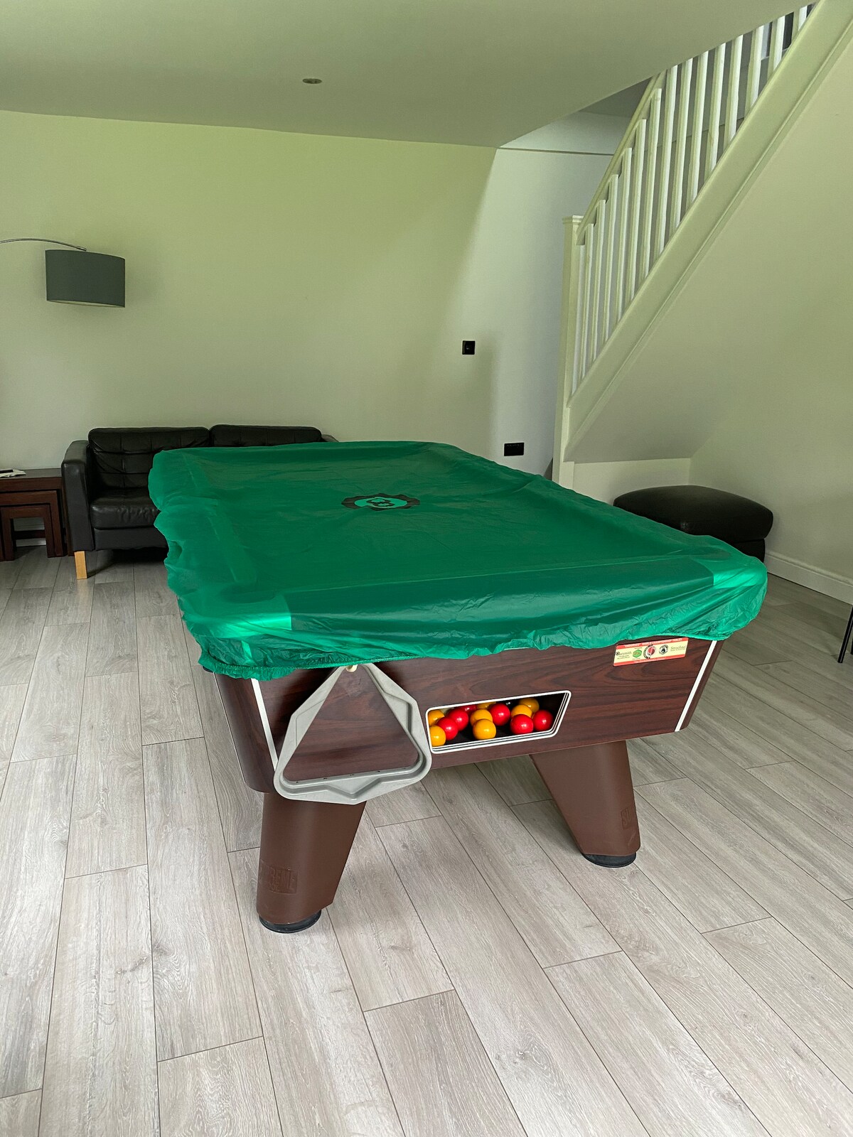 A pool table is covered with a green protective sheet, positioned in a spacious room with light wood flooring. Nearby, a black sofa is visible, along with a small table. Natural light illuminates the area, enhanced by the minimalist decor and the staircase in the background.