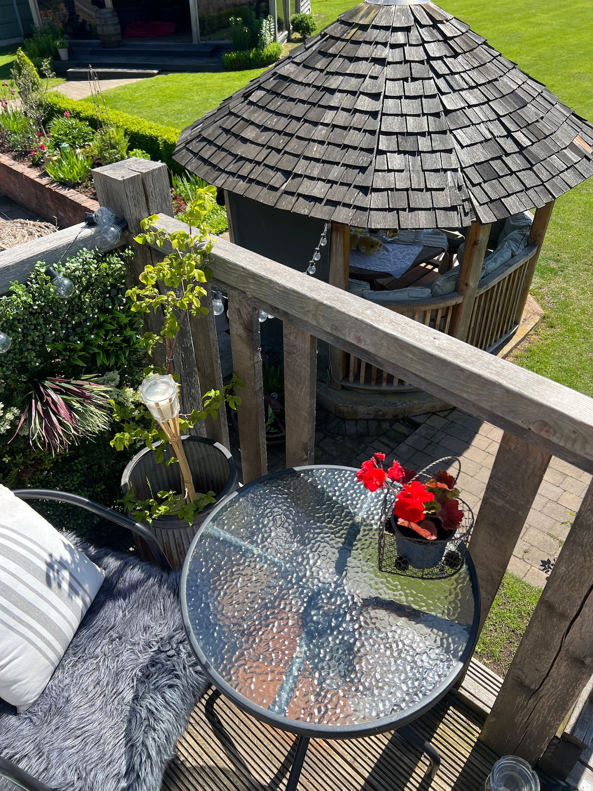 A small round table with a textured glass top is set on a wooden balcony, accompanied by a chair with a cozy cushion. A potted red flower adds a touch of color. In the background, a circular Cedar Pavilion is visible, surrounded by well-manicured grass and greenery.