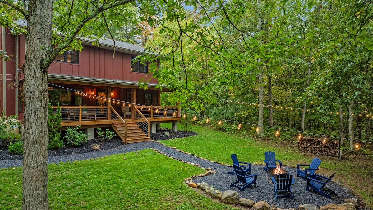 A serene cabin exterior is framed by lush greenery, featuring a wooden deck and steps leading to a welcoming entrance. Four blue adirondack chairs are arranged around a fire pit, highlighted by soft outdoor lights strung above, creating an inviting outdoor gathering space.