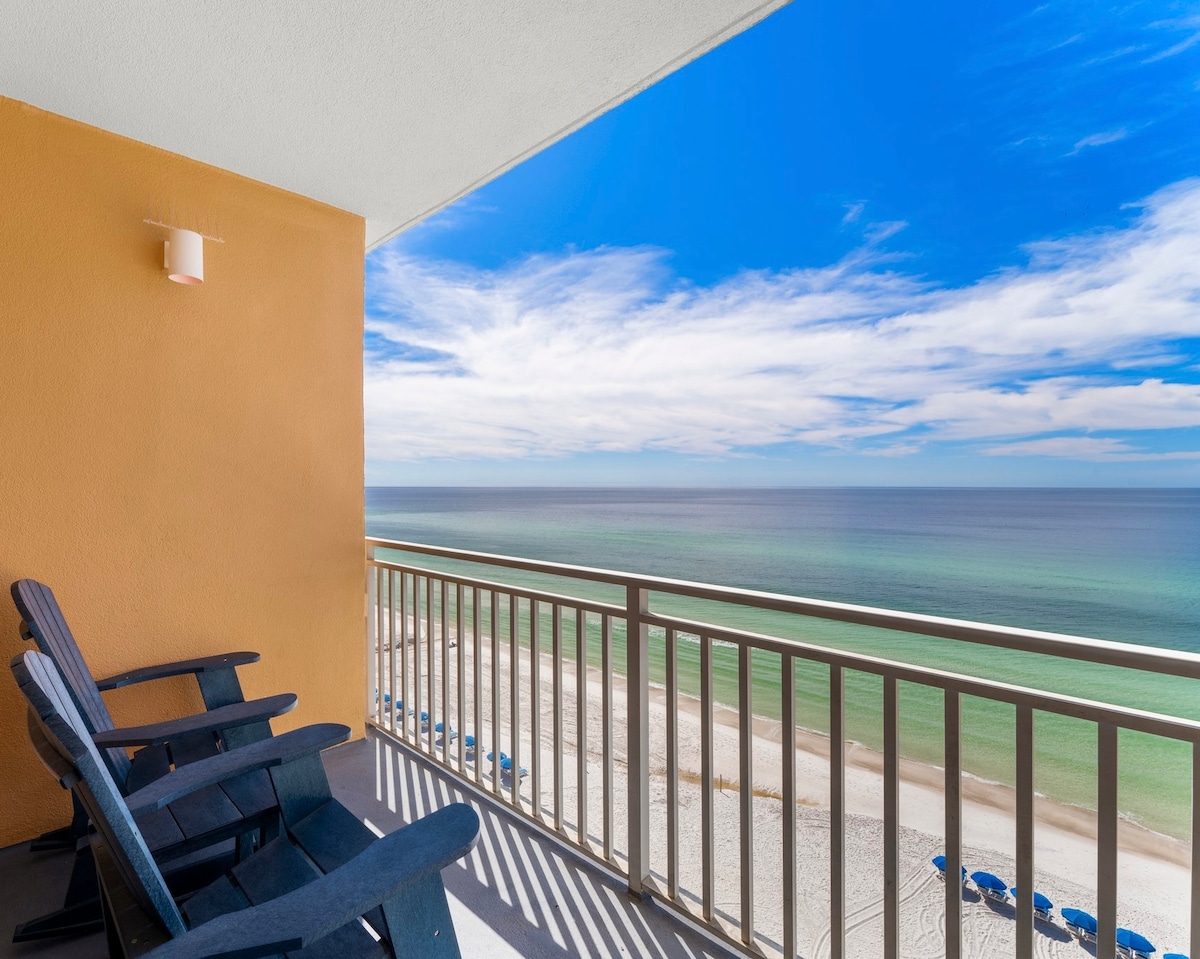 A spacious balcony showcases expansive beach views, with a calm ocean and sandy shore in the distance. Two Adirondack chairs are placed on the balcony, inviting relaxation while enjoying the scenic surroundings and bright blue sky.