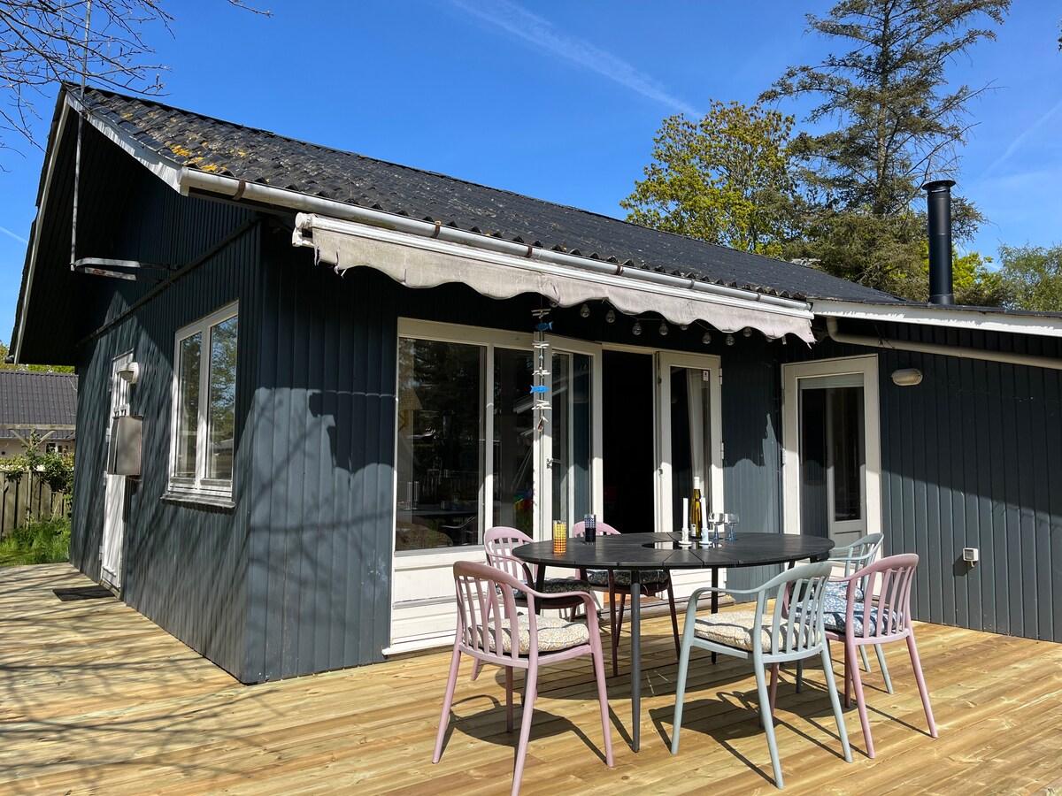 A spacious outdoor terrace features a large wooden deck with a dining table surrounded by colorful chairs. A retractable awning provides shade, while trees and greenery are visible in the background. A door leads to the interior, creating a seamless flow between indoor and outdoor spaces.