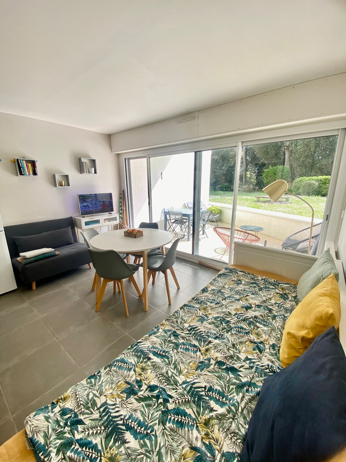 A bright living area features a spacious convertible sofa bed and a circular dining table surrounded by four chairs. Large sliding glass doors provide a view of the outdoor space, which includes a patio with seating. Natural light enhances the room's fresh atmosphere.