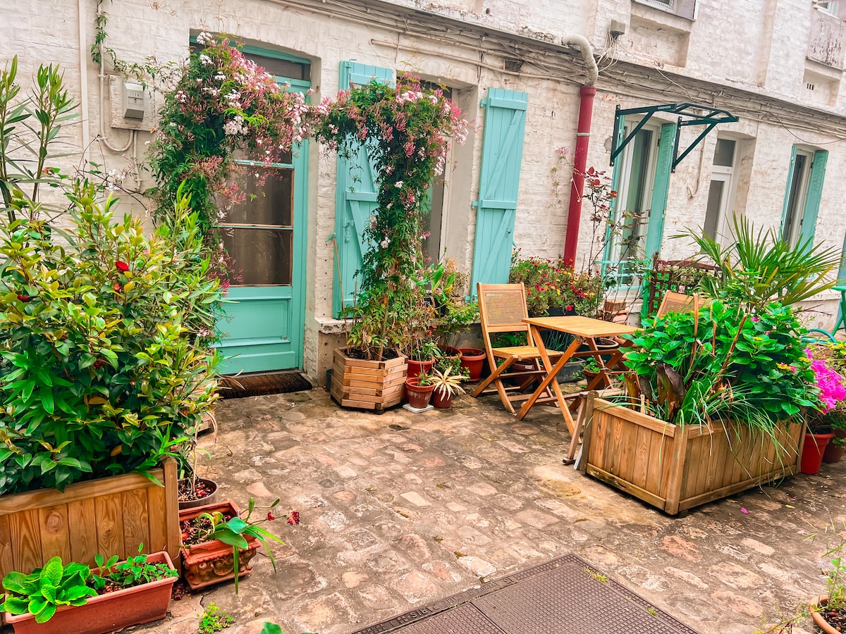An outdoor space is framed by greenery and flower-filled planters. A small wooden table and folding chairs are situated in the center, surrounded by vibrant plants. The entrance features a soft aqua door, adding a touch of color to the charming courtyard.