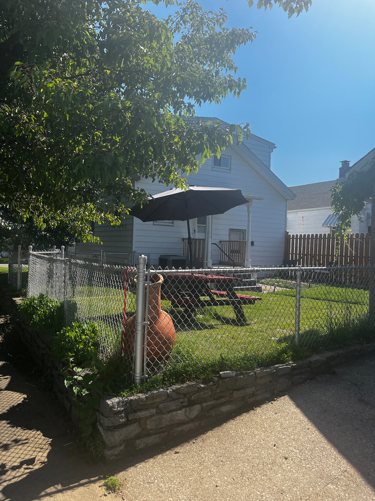 A well-maintained backyard is enclosed by a chain-link fence, featuring a grassy area with a picnic table shaded by a large black umbrella. A stone border adds structure to the space, while a gas grill is positioned nearby, ready for outdoor cooking.