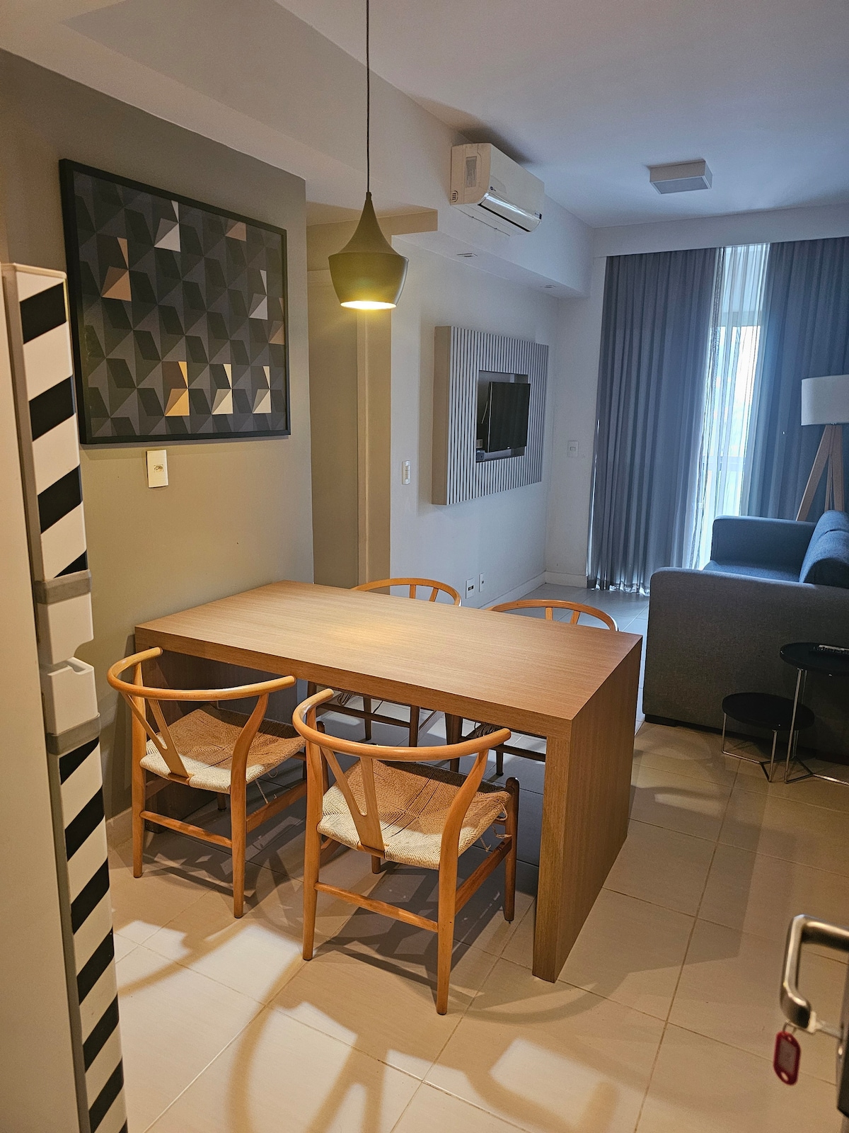 A modern dining area is featured with a wooden table and two simplistic chairs, complemented by a geometric wall art piece. A cozy lounge area can be seen in the background with a comfortable gray sofa and a mounted television, illuminated by natural light from large windows.