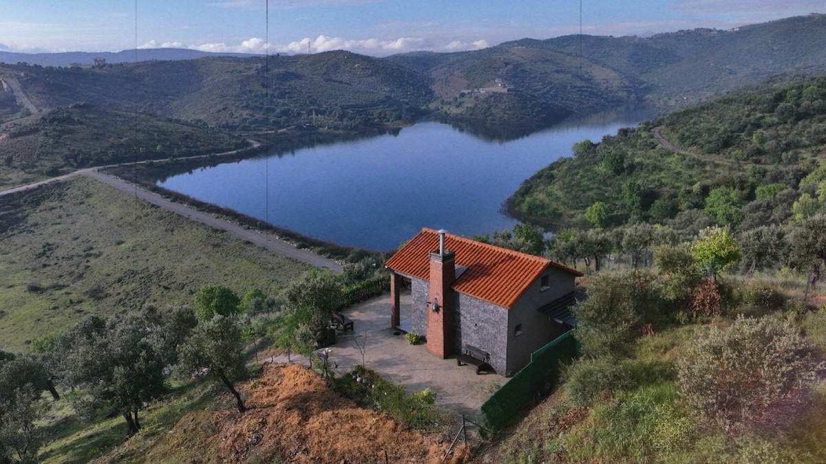 A mountain house is nestled in a verdant landscape, overlooking a tranquil lake. Surrounding hills are framed by greenery, while the structure features a red-tiled roof and a chimney. A wide patio area extends from the home, providing outdoor space with scenic views.