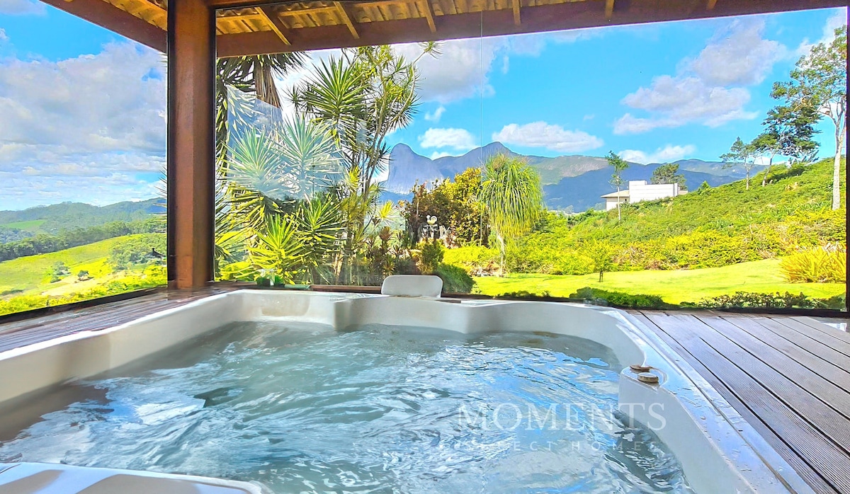 A private jacuzzi is positioned on a wooden deck, surrounded by large windows that reveal expansive views of lush greenery and distant mountains. Bright sunlight enhances the serene atmosphere, inviting relaxation amidst nature.
