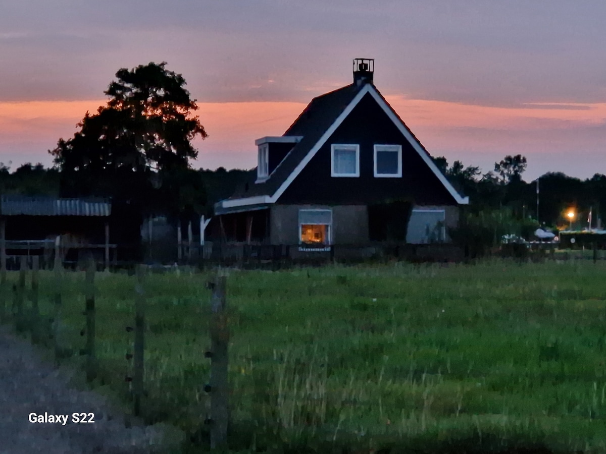 A charming house with a dark exterior is set against a serene twilight sky. The structure features a gabled roof and a prominent cupola, with a warm light visible from a window. Surrounding fields and a fenced area enhance the peaceful rural setting.