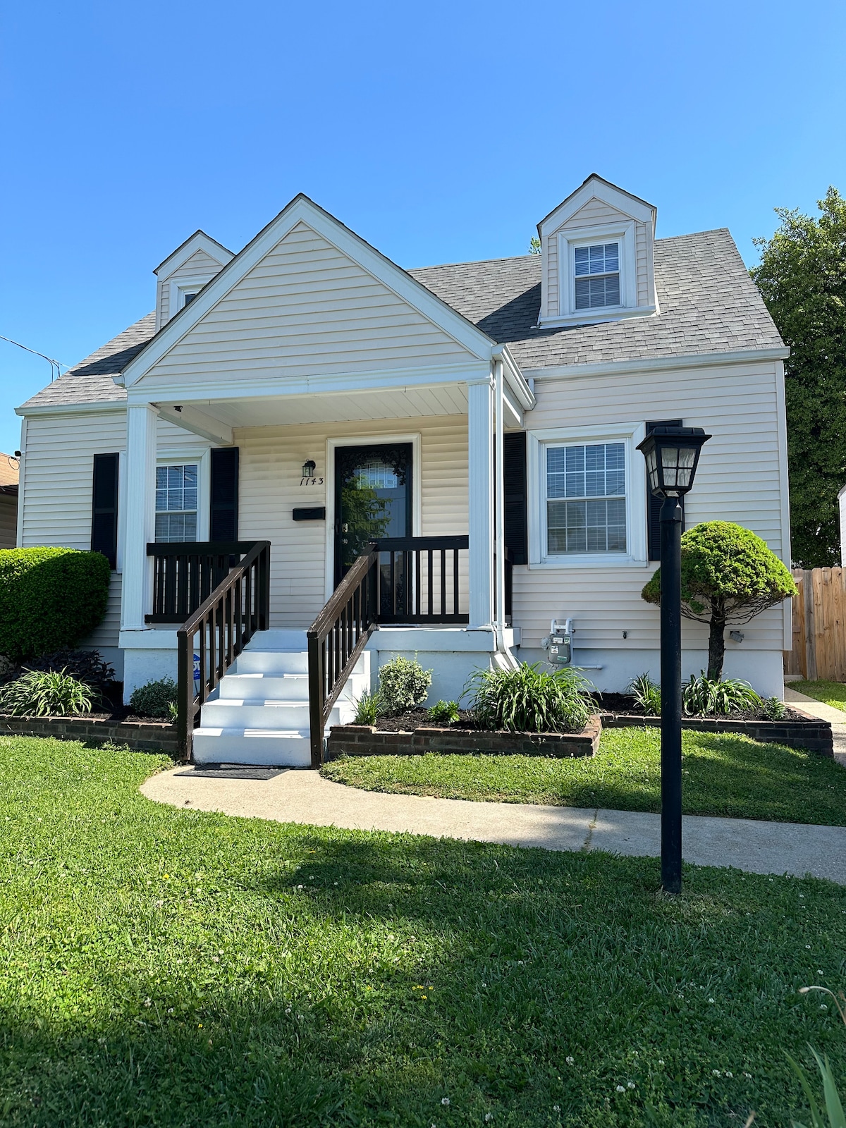 A charming two-story home is depicted with a light-colored exterior and a sloped roof. The front porch features white columns and black railing. Lush green landscaping surrounds the entryway, and a lamp post stands nearby, complementing the well-maintained front yard.