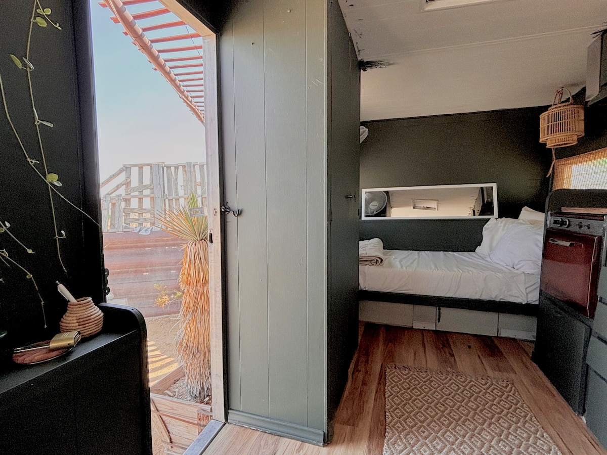 A compact living space is shown, featuring a spacious bed with a layered white bedding arrangement. A mirror reflects the area, and a wooden rug complements the light wood flooring. Natural light enters through an open door, highlighting a simple and functional design.