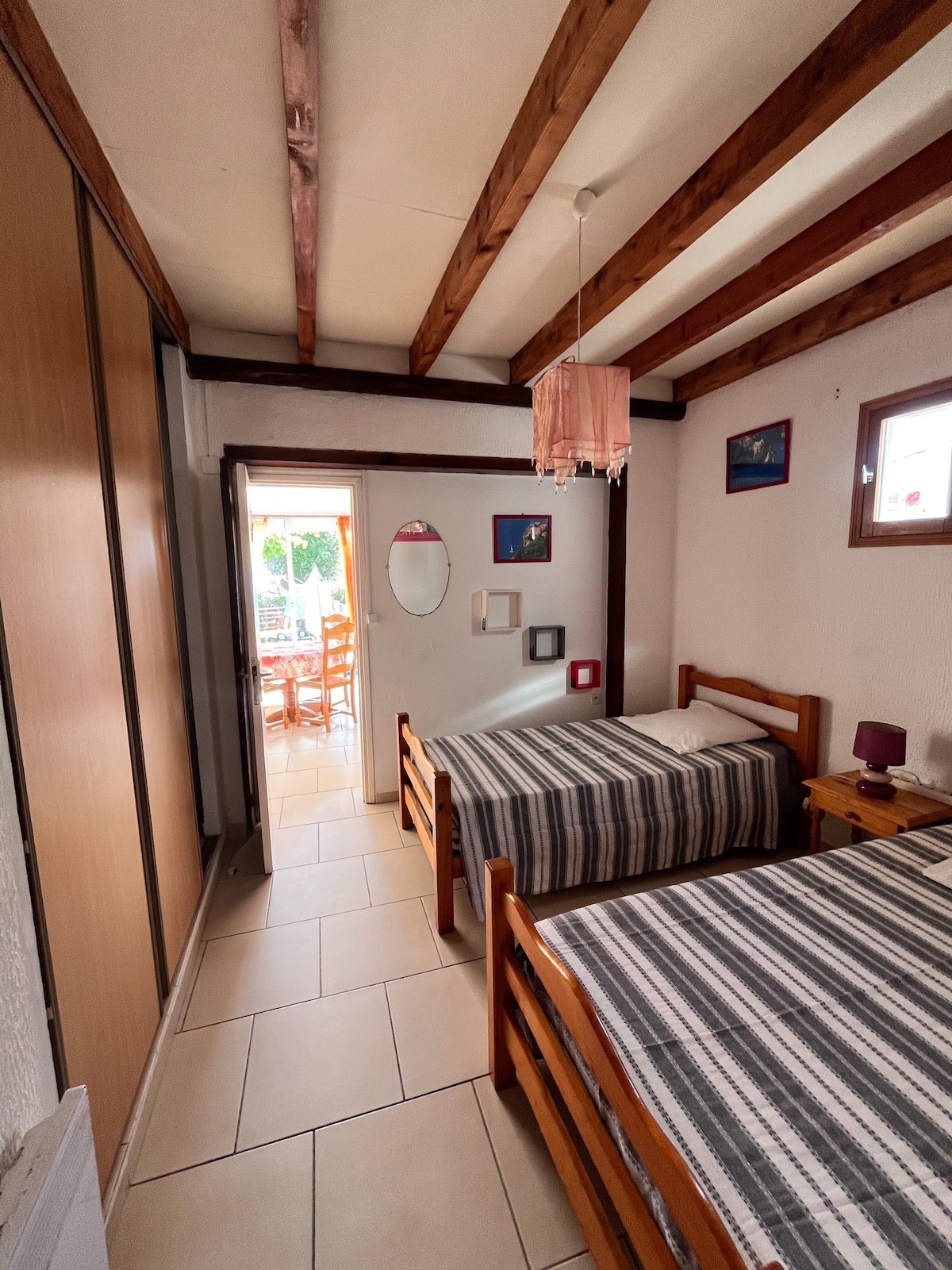 The room features two individual beds with striped bedding and wooden frames. A closet spans one wall, while a doorway leads to a patio area with outdoor seating. Natural light filters in through a window, complemented by a simple hanging light fixture.