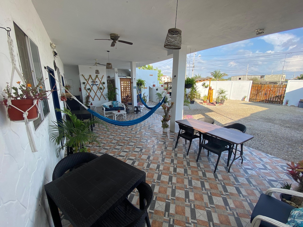 An outdoor patio area features a cozy seating arrangement with black chairs and tables. A hammock is suspended nearby, surrounded by plants. The tiled floor showcases a colorful geometric pattern, while light filters in through the open spaces, enhancing a welcoming atmosphere.
