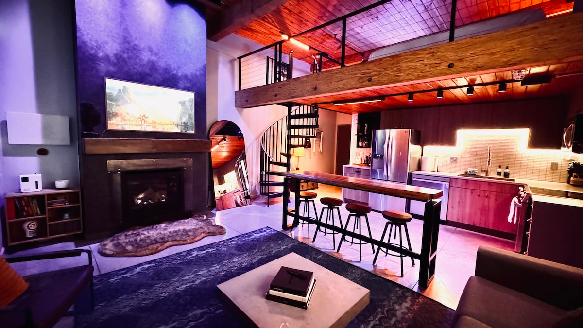 An inviting loft space features a modern fireplace beneath a large smart TV. A spiral staircase leads to a second floor. The kitchen showcases stainless steel appliances and a wooden island with seating for four. Ambient lighting enhances the overall design, creating a cozy atmosphere.