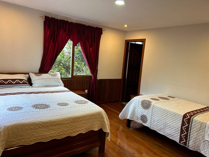 Habitación principal con cama matrimonial y cama individual para 3 huéspedes con clóset y baño con ducha caliente con vista a los jardines laterales y al frente, vista al río Upano y las montañas