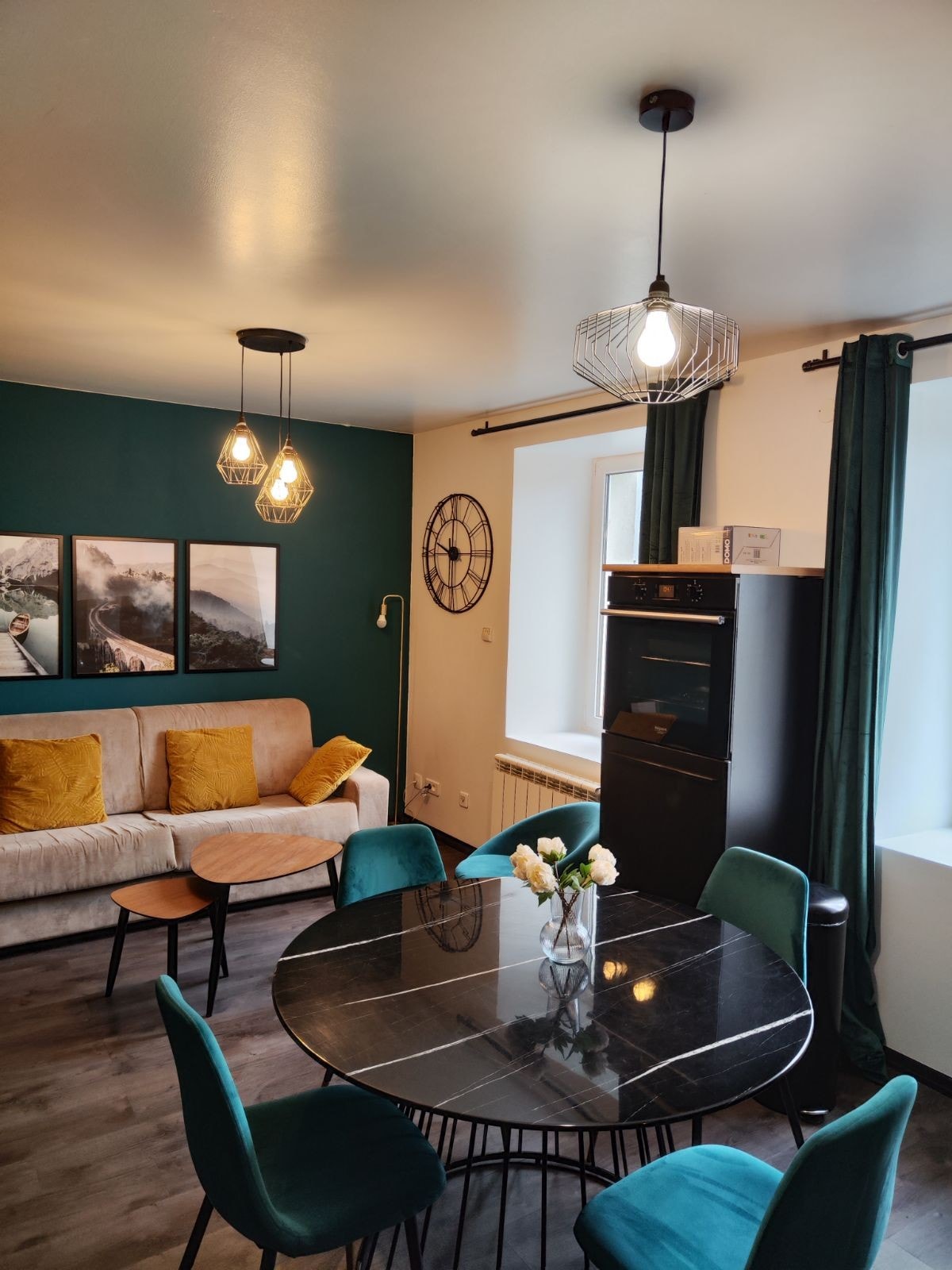 A cozy living space features a light-colored sofa with plush cushions, positioned against a dark green accent wall. A round, black dining table is surrounded by teal chairs, with a vase of white flowers as the centerpiece. Decorative artwork adorns the walls, and large windows allow natural light.