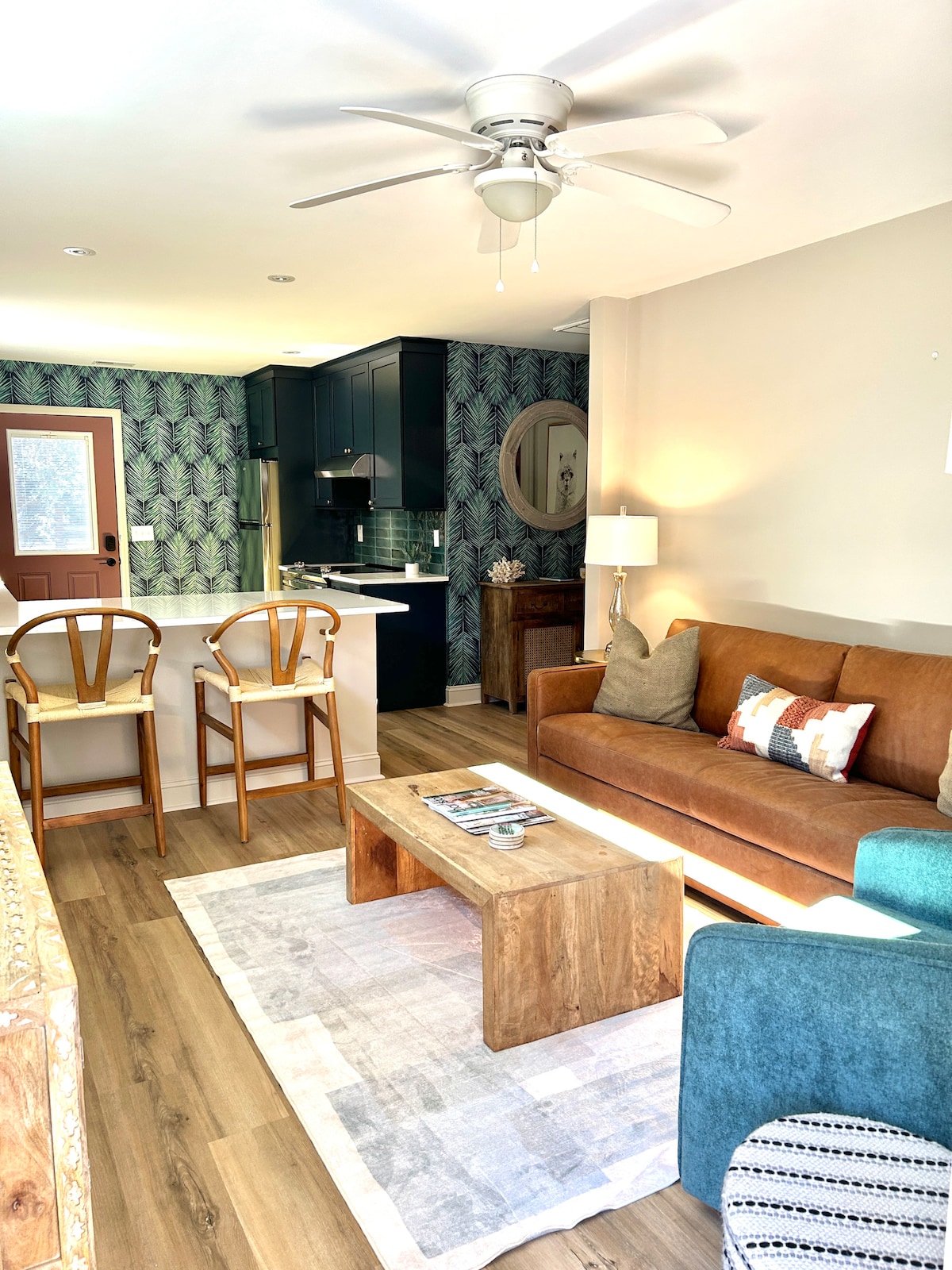A bright living area is depicted with a brown sofa, accented by decorative pillows and a wooden coffee table. Natural light enters through a door, enhancing the green floral wallpaper and wooden flooring. Two bar stools are positioned at the kitchen counter in the background.