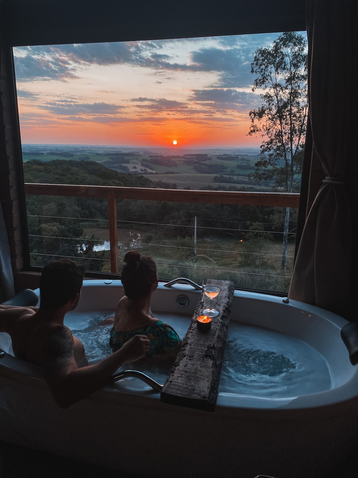 A spacious hot tub is positioned by large windows, offering a view of a vibrant sunset over rolling hills. A wooden ledge holds two drinks, while two guests relax comfortably in the warm water, framed by soft curtains.