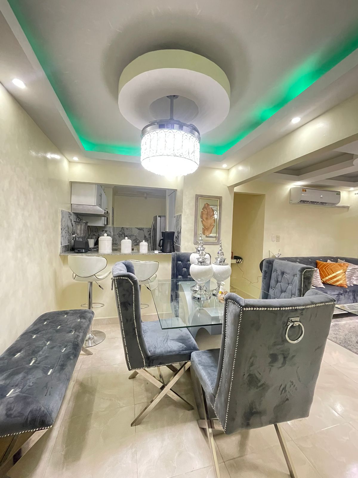 A dining area features a glass table surrounded by six velvet chairs with chrome accents. An elegant chandelier hangs from the ceiling, complemented by a subtle green lighting effect. A plush sofa is positioned in the background, creating a welcoming space for gatherings.