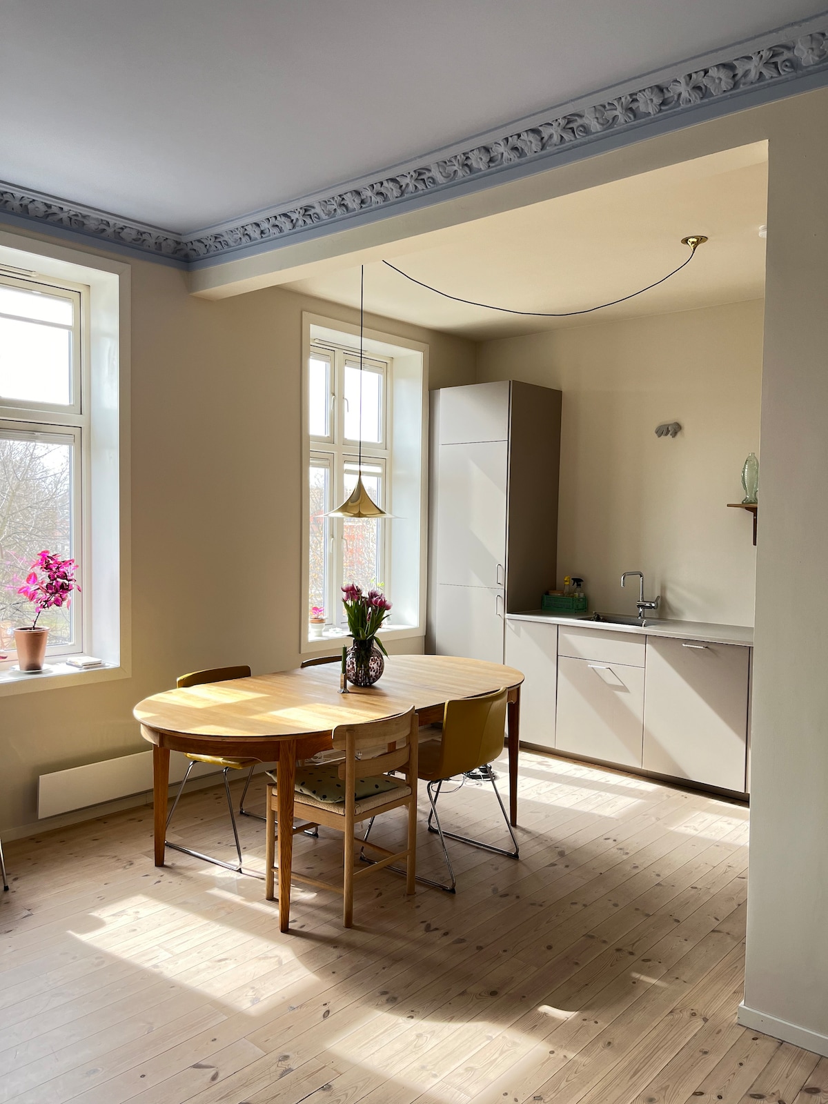 A bright kitchen area features a round wooden table surrounded by four chairs. Two large windows allow natural light to pour in, highlighting the neutral walls and cabinetry. A small sink is visible, alongside modern kitchen appliances, creating a functional space for dining and cooking.