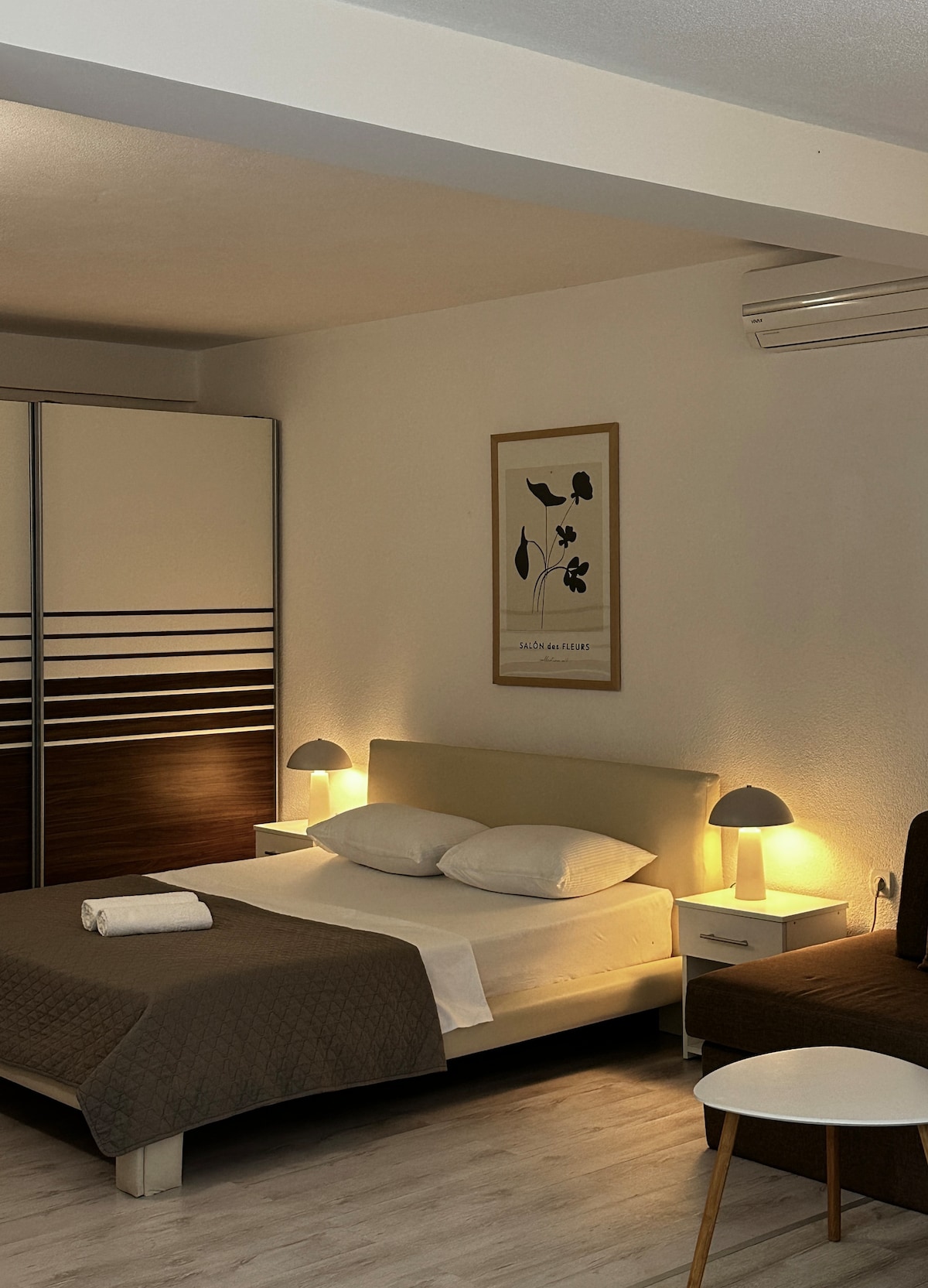 A well-lit studio apartment features a spacious bed with neutral bedding. Soft bedside lamps provide gentle illumination, and a wardrobe is positioned against the wall. A small table and chair are present, complementing the cozy atmosphere of the space.