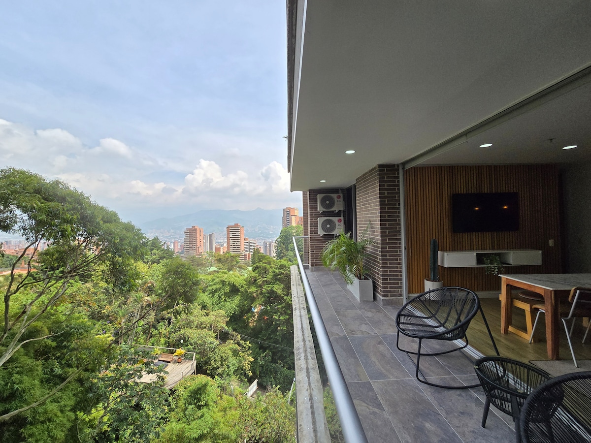 A spacious balcony is featured with views of the city skyline and surrounding greenery. Modern outdoor seating accompanies a dining table made of wood. Air conditioning units are visible along the building's exterior, blending with the natural scenery.
