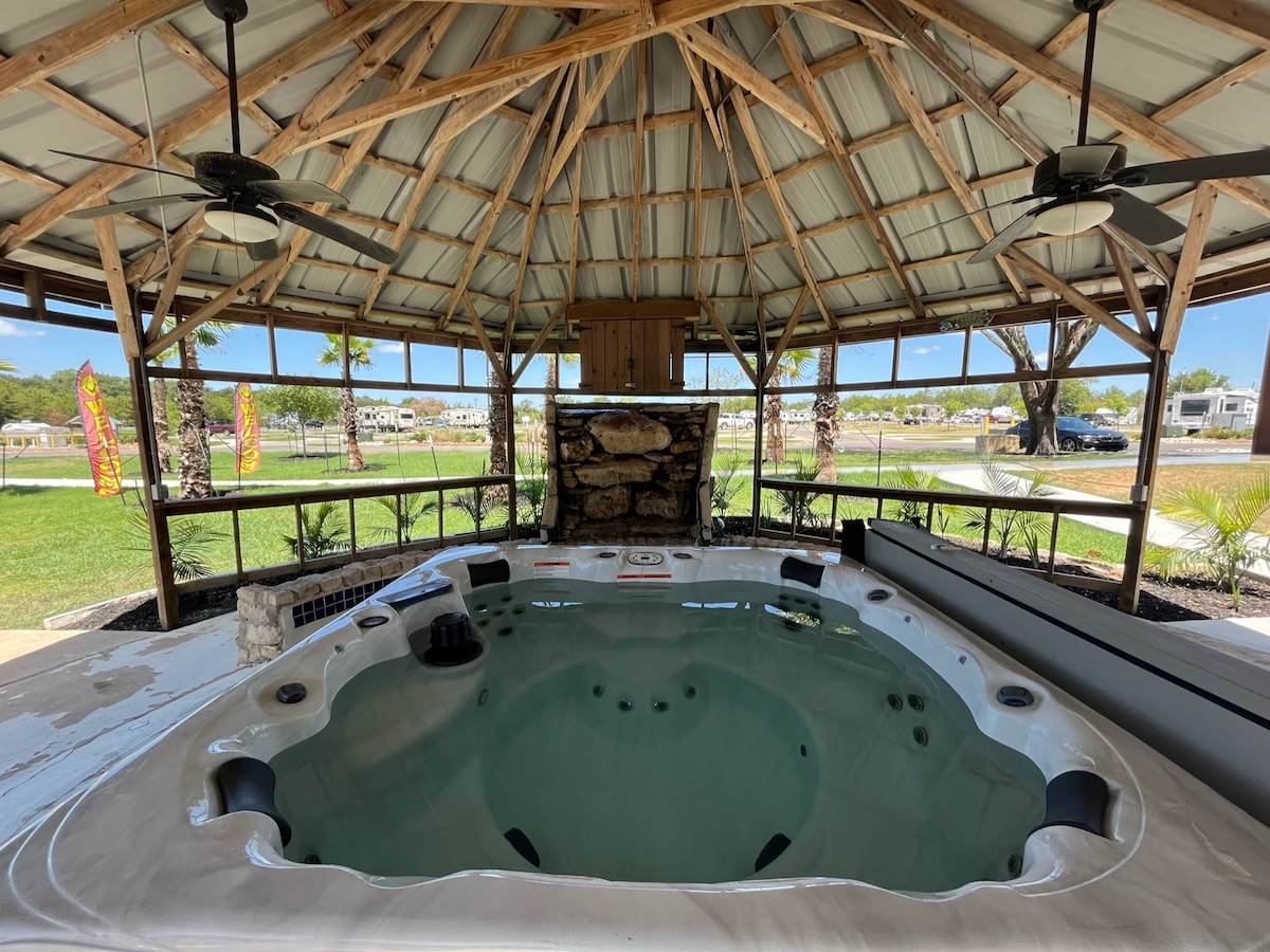A sheltered area features a hot tub, surrounded by a rugged stone wall and ceiling fans for comfort. Natural light filters through the open design, illuminating the tub's interior, while grassy grounds extend beyond the structure.