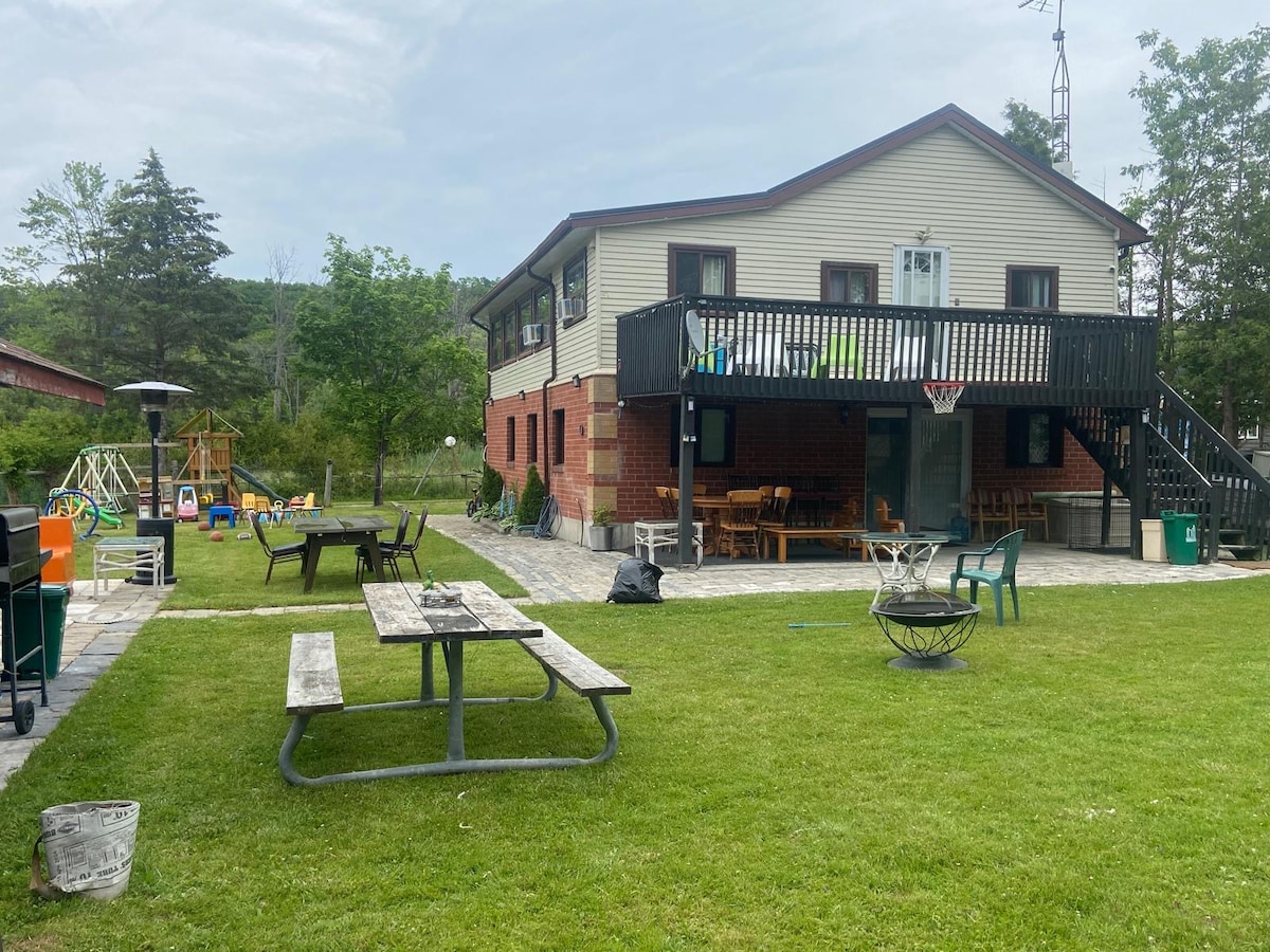The exterior of a two-story home is visible, featuring a spacious balcony and patio area. A picnic table and a fire pit are positioned on the well-maintained lawn, surrounded by seating options. Play equipment is present, with lush greenery and trees enhancing the outdoor space.