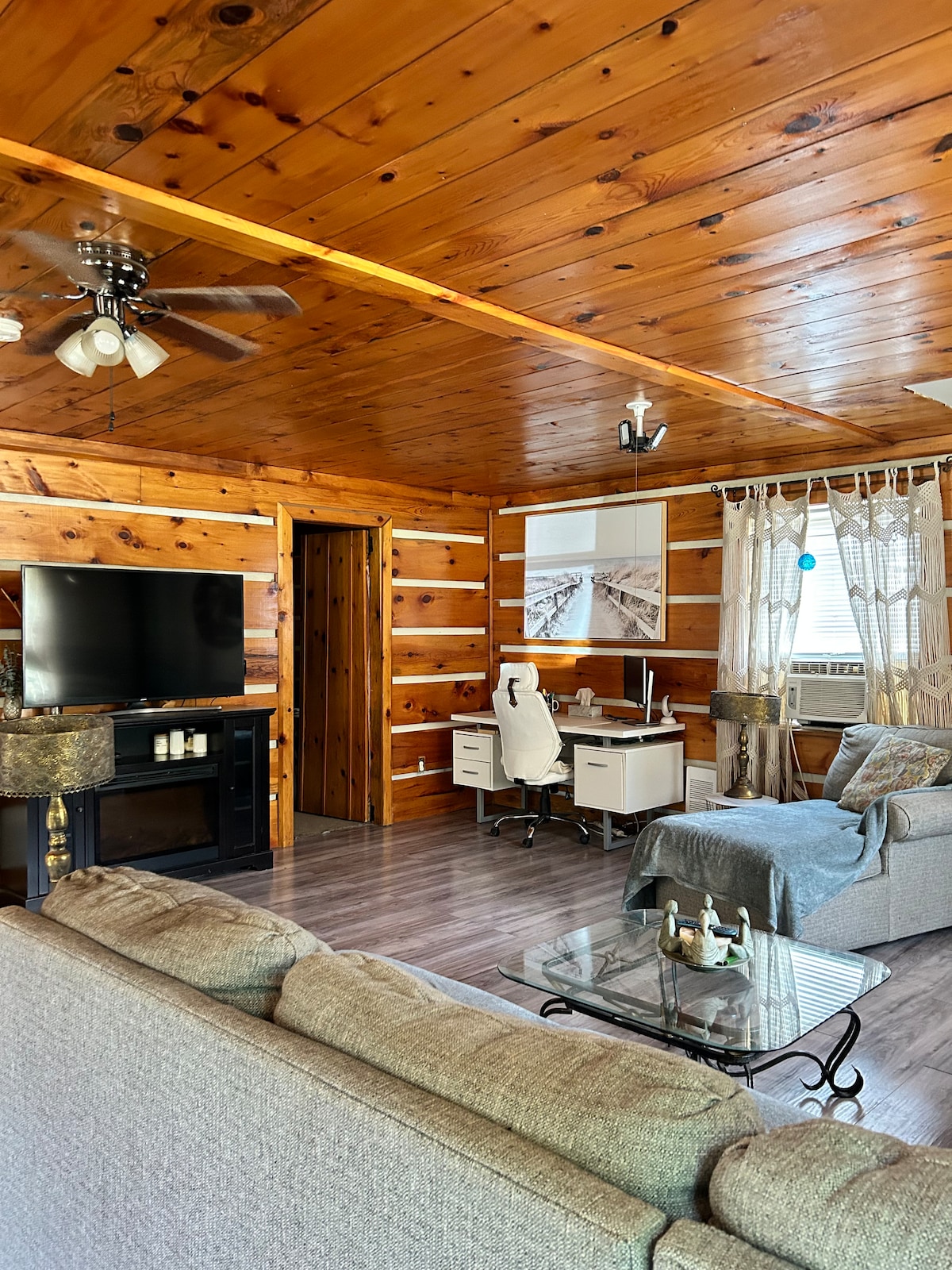 The living area features wooden paneling and a ceiling fan. A plush gray sofa is positioned near a glass coffee table, with a television mounted on the wall. A well-lit workspace with a white desk and chair is present, along with natural light filtering through sheer curtains.